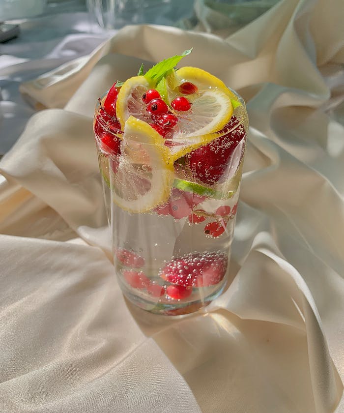 Clear glass of citrus berry detox water with lemon, pomegranate, and mint on satin fabric.